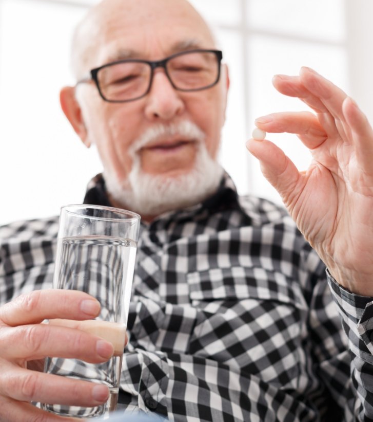 An old man holding a pill between his fingers and a glass of water in his other hand